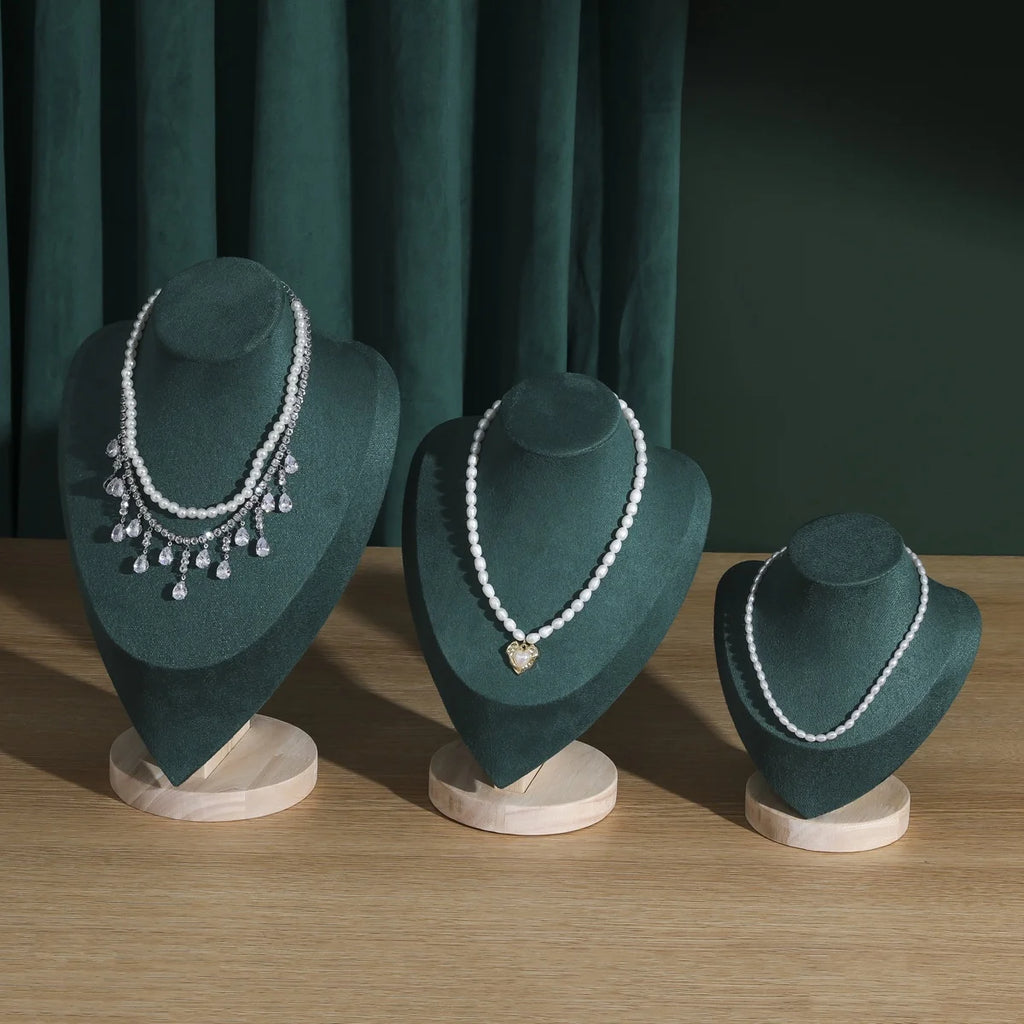 Buste en bois Nesty - Trois présentoirs en velours vert sur table en bois avec colliers de perles, devant un rideau vert