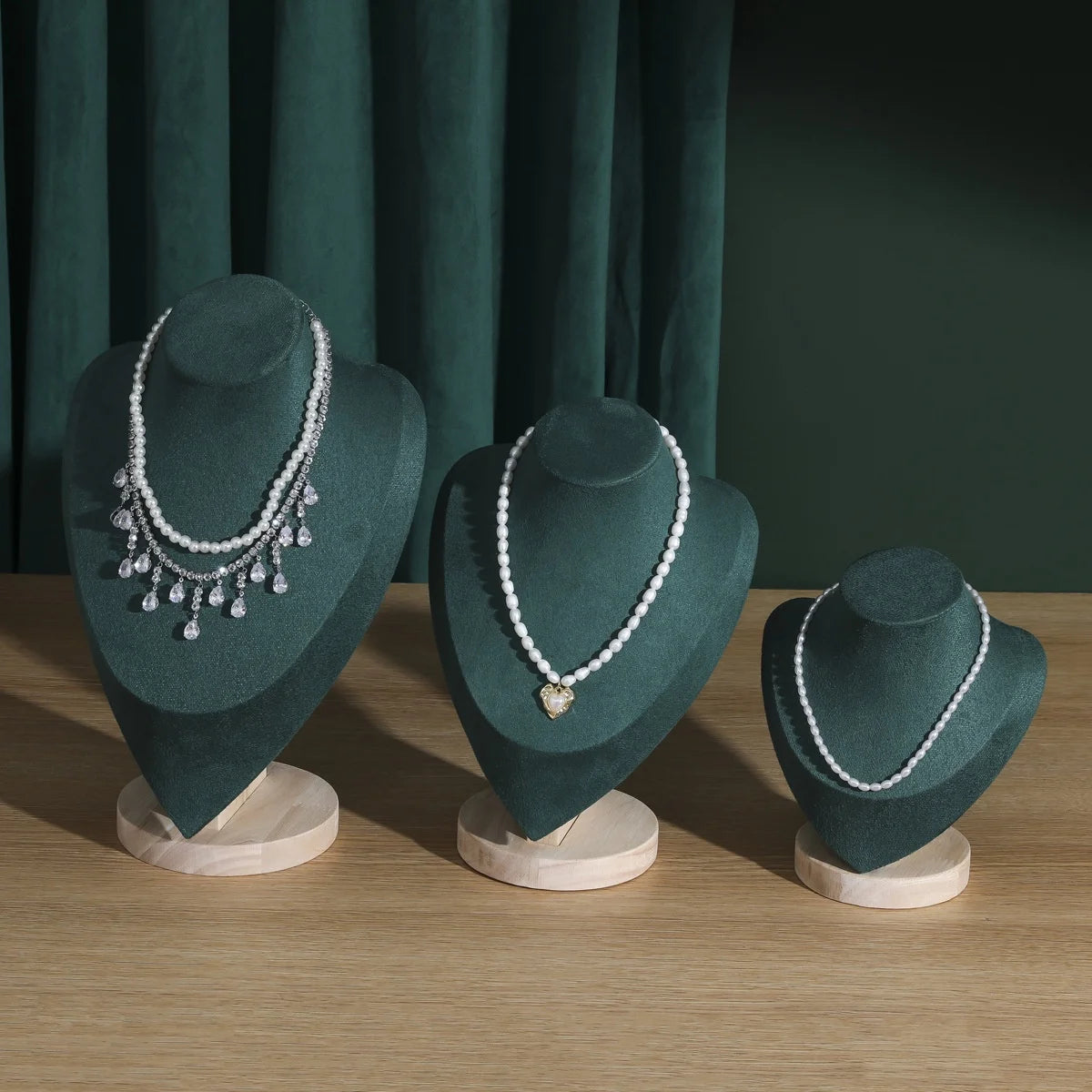 Buste en bois Nesty - Trois présentoirs en velours vert sur table en bois avec colliers de perles, devant un rideau vert