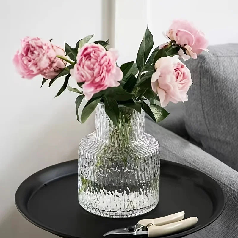 Vase en verre soufflé texturé glacier, 8 cm de diamètre Nesty, posé sur un plateau noir, garni de pivoines roses, canapé gris en arrière-plan