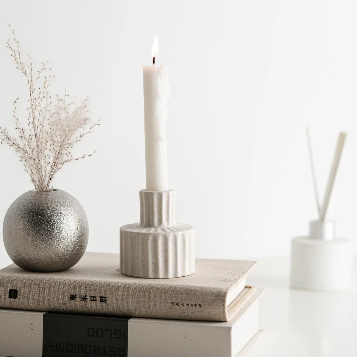 Bougeoir céramique nordique godronné blanc, bougie blanche allumée, sur pile de livres avec vase en métal argenté et bâtons parfumés - Nesty