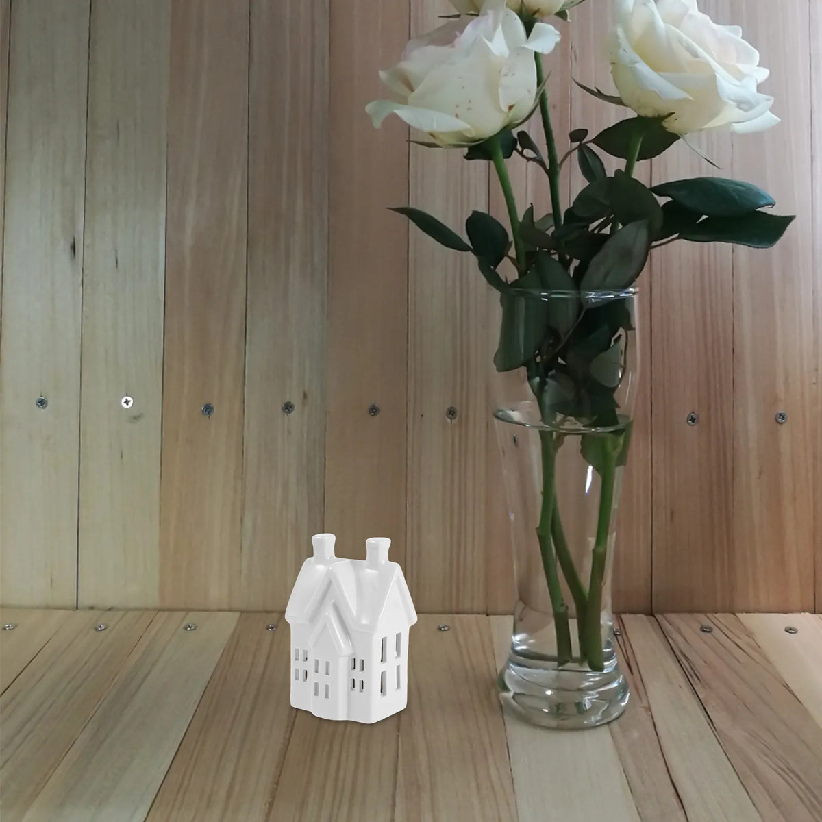 Photophore maisonnette ajourée en céramique Nesty - Maisonnette posée sur des lattes de bois à côté d'un vase de roses blanches