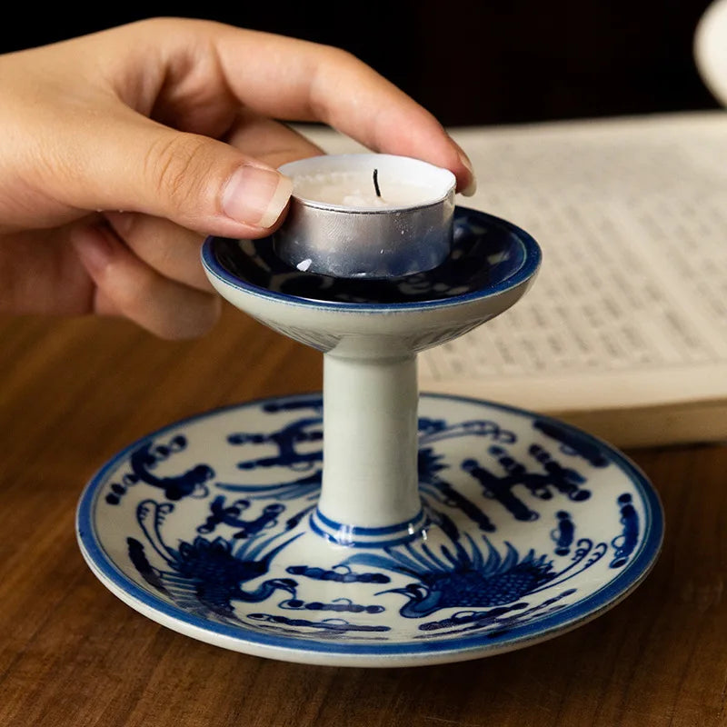 Main posant une bougie chauffe-plat sur un bougeoir en porcelaine peinte bleu et blanc motif oiseaux, devant un livre ouvert - Nesty