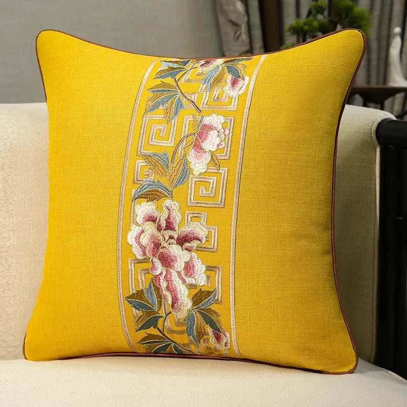 Housse de coussin en coton et lin, broderie florale classique Nesty, housse de coussin jaune moutarde avec bande brodée de fleurs roses et motif grec doré, posée sur fauteuil
