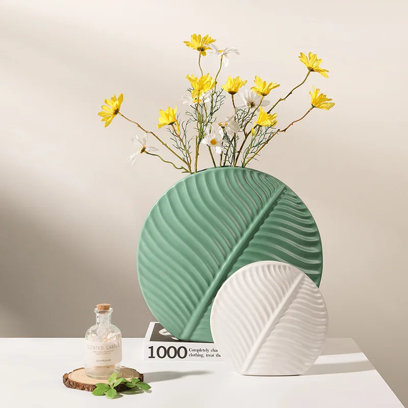 Vase céramique de style européen, motif feuille - Nesty - Grand modèle vert sauge et petit modèle blanc en forme de feuille, avec marguerites jaunes et blanches, sur une table près d'une bougie parfumée