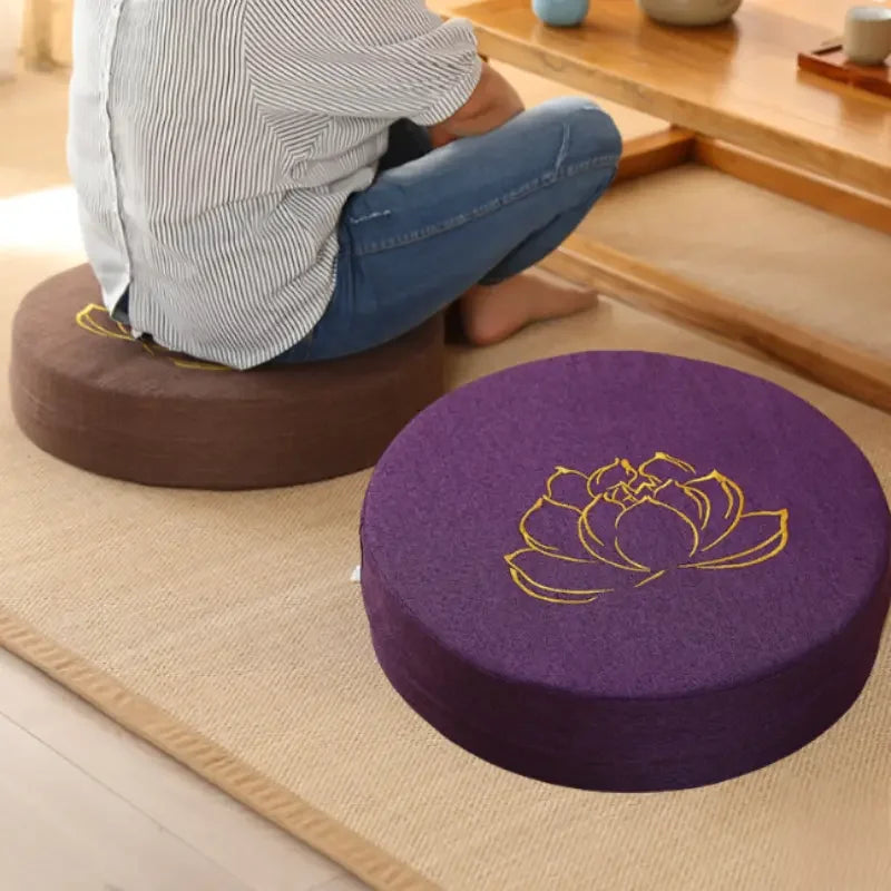 Coussin de méditation en lin, texture lotus, modèle compact Nesty, coussin rond de méditation violet brodé d'un lotus doré, posé sur tatami à côté d'un coussin brun sur lequel une personne est assise en tailleur