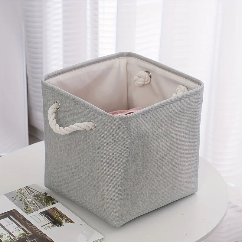 Boîte de rangement pliable en lin pour vêtements Nesty, boîte grise posée sur une table ronde blanche avec magazine, devant un rideau