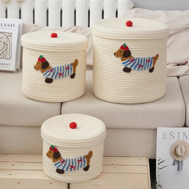 Panier de rangement pliable en corde de coton, grande capacité Nesty, trois tailles avec patch brodé de teckel et pompon rouge, posées sur un canapé, radiateur en fond