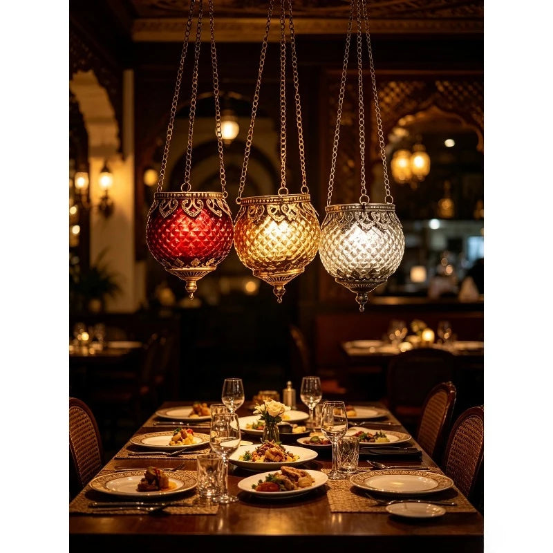 Trio de photophores suspendus en verre Nesty - Trois lanternes rouge, dorée et argentée allumées, suspendues au-dessus d'une table dressée dans un restaurant