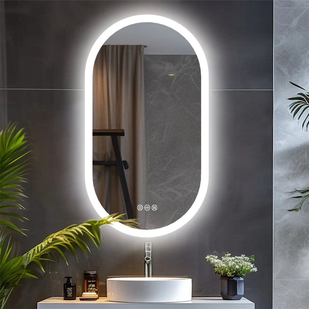 Miroir de salle de bain ovale LED Nesty - Allumé dans une salle de bain en pierre grise, reflet d'un rideau et d'une plante
