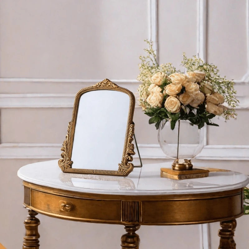 Miroir de coiffeuse doré — cadre en bois ancien - Nesty, posé sur une table dorée à plateau marbré, à côté d'un bouquet de roses crème