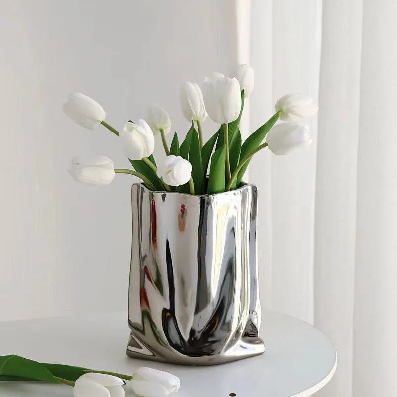 Vase en céramique argentée, forme sac plissé Nesty, effet froissé argenté garni de tulipes blanches, sur table blanche avec rideau