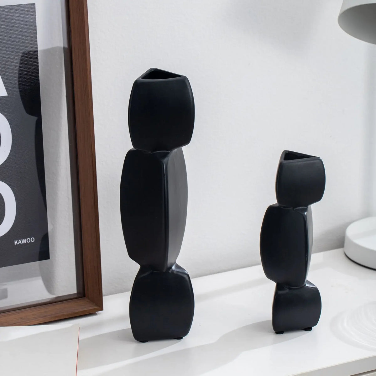 Vase noir en céramique Nesty - Gros plan sur deux modèles noirs empilés en galets, près d'une affiche encadrée