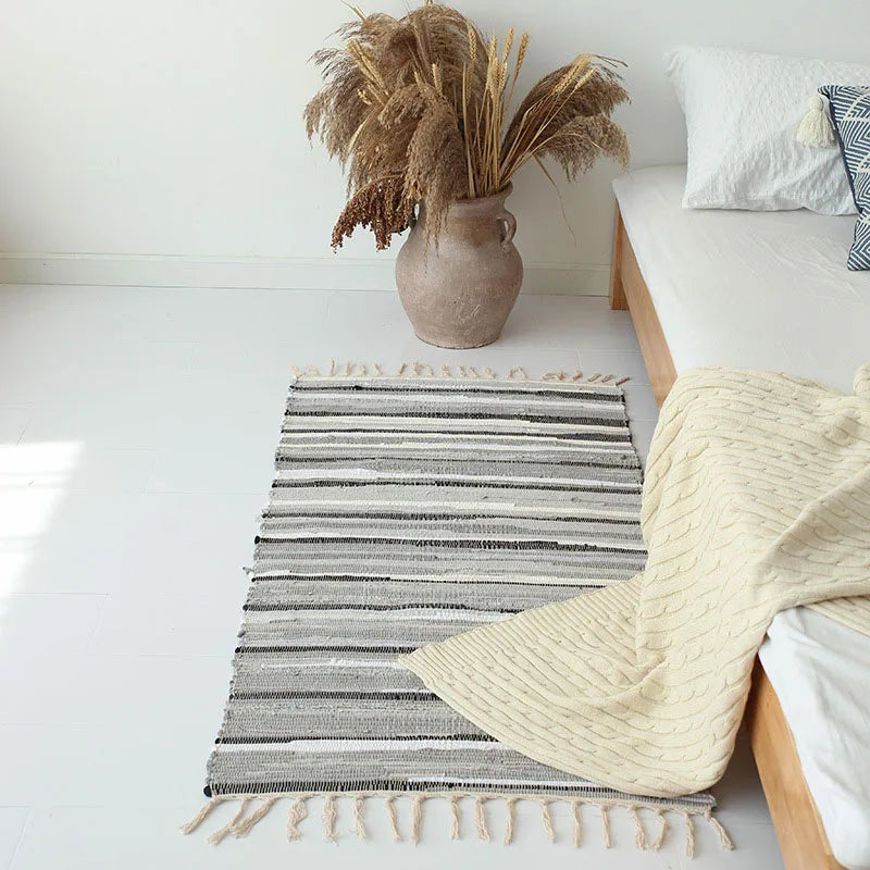 Tapis à franges tissé main — motif rayé - Nesty, variante à rayures grises, noires et blanches avec franges, posée près d'un vase de graminées séchées