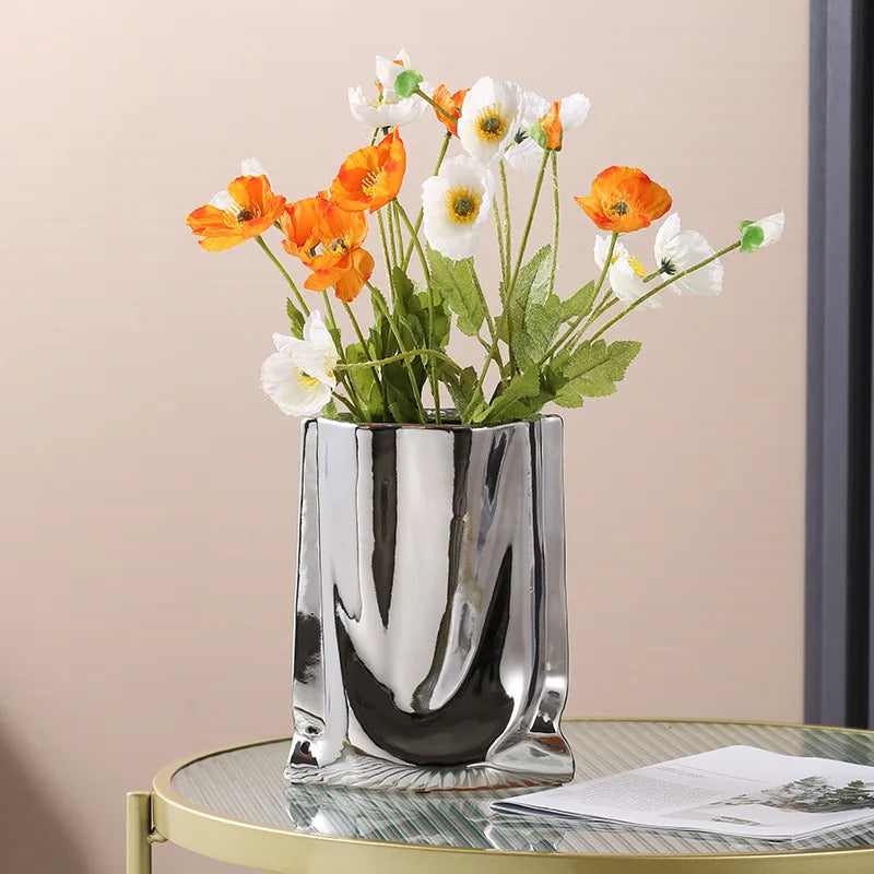 Vase en céramique argentée, forme sac plissé Nesty, effet froissé argenté garni de coquelicots orange et blancs, sur table ronde dorée