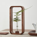 Vase soliflore en bois et verre Nesty - Cadre rectangulaire en noyer et tube en verre garni d'une fougère, sur une table